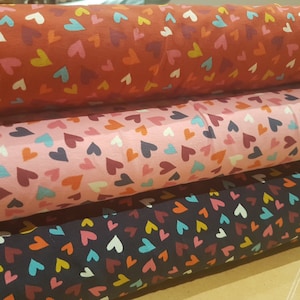 May include: Three rolls of fabric with a repeating pattern of colorful hearts. The fabric is available in red, pink, and navy blue.
