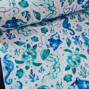 May include: White fabric with a repeating pattern of marine life in shades of blue and teal. The design includes seahorses, jellyfish, sea turtles, stingrays, seashells, and coral. The fabric is rolled, showing the pattern's repeat.