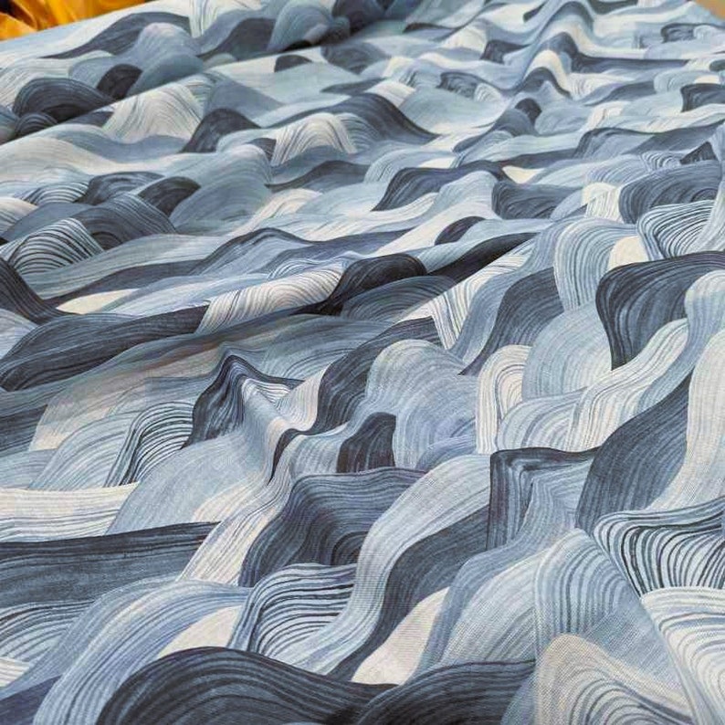 May include: A close-up of a fabric with a flowing, wave-like pattern in shades of blue and white. The design resembles a stylized landscape of rolling hills or ocean waves, creating a textured and dynamic visual effect. The fabric appears to be soft and pliable.