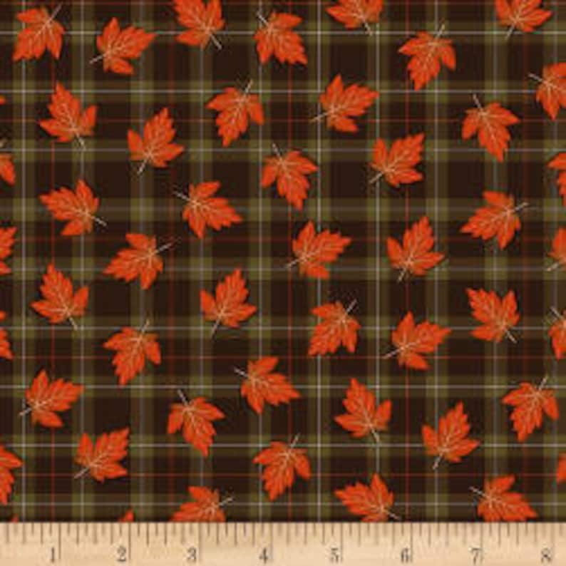 May include: Autumn-themed fabric featuring a plaid pattern in shades of brown, green, and red. Scattered across the plaid are orange maple leaves. A wooden ruler with inch markings is at the bottom.