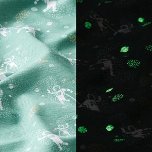 Puede incluir: Una tela verde con un estampado blanco de astronautas y estrellas. La tela brilla en la oscuridad, revelando un estampado verde de astronautas y estrellas.