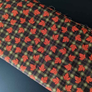 May include: Fabric with a repeating pattern of red maple leaves on a dark brown plaid background with green and white lines. The fabric is folded, showing the pattern at an angle.