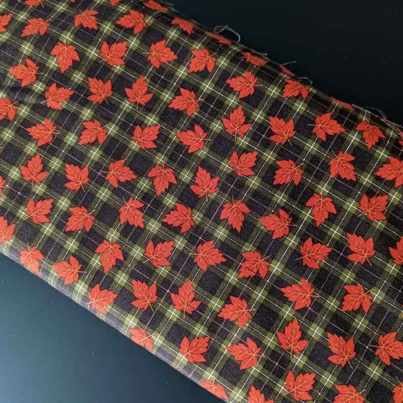 May include: Fabric with a repeating pattern of red maple leaves on a dark brown plaid background with green and white lines. The fabric is folded, showing the pattern at an angle.