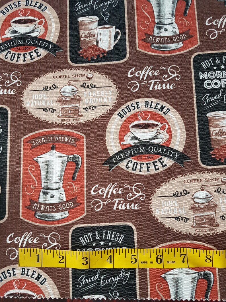 May include: A brown fabric with a repeating pattern of coffee-themed illustrations. The illustrations include coffee cups, coffee pots, coffee beans, and text that says "House Blend", "Premium Quality Coffee", "Coffee & Time", "Hot & Fresh Morning Coffee", "Served Everyday", "Coffee Shop", "Locally Brewed", "Always Good", and "100% Natural". A yellow measuring tape is placed across the bottom of the fabric.