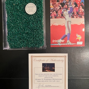 May include: A certificate of authenticity for a piece of turf from the Hubert H. Humphrey Metrodome, signed by Guy R. Nelson, President, Elite Sports Promotions Agency. The certificate includes a photo of the Metrodome at night. The certificate is accompanied by a small plastic container with a piece of green turf inside and a football card of Cris Carter, a former Minnesota Vikings player.