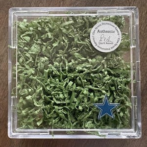 May include: A clear plastic case filled with green shredded paper, a blue star, and a circular sticker that reads "Authentic" and "Elite Sports Promotions Agency". The case is on a wooden surface.