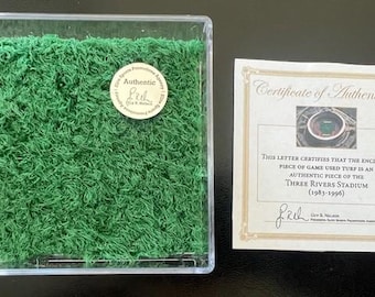 May include: A section of green turf presented in a clear acrylic case, alongside a certificate of authenticity. The certificate confirms the turf is from Three Rivers Stadium (1983-1996). A round sticker on the case reads "Authentic".