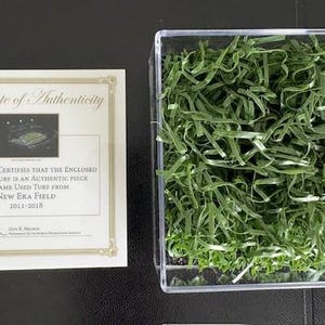 May include: A certificate of authenticity for a piece of game-used turf from New Era Field, 2011-2018. The certificate is signed and includes a small photo of the field. The turf is displayed in a clear plastic box with green shredded paper.