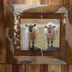 May include: A rustic wooden frame with a white cowhide mat surrounds a photograph of two white cows standing in a field. The photograph is signed "WF" in the bottom right corner.