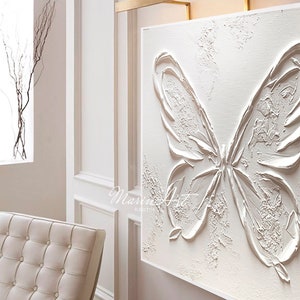 Op de afbeelding: Witte textuurverf op canvas van een vlinder met een gouden metalen frame en een lamp erboven. Het schilderij is getiteld "Marin Art".