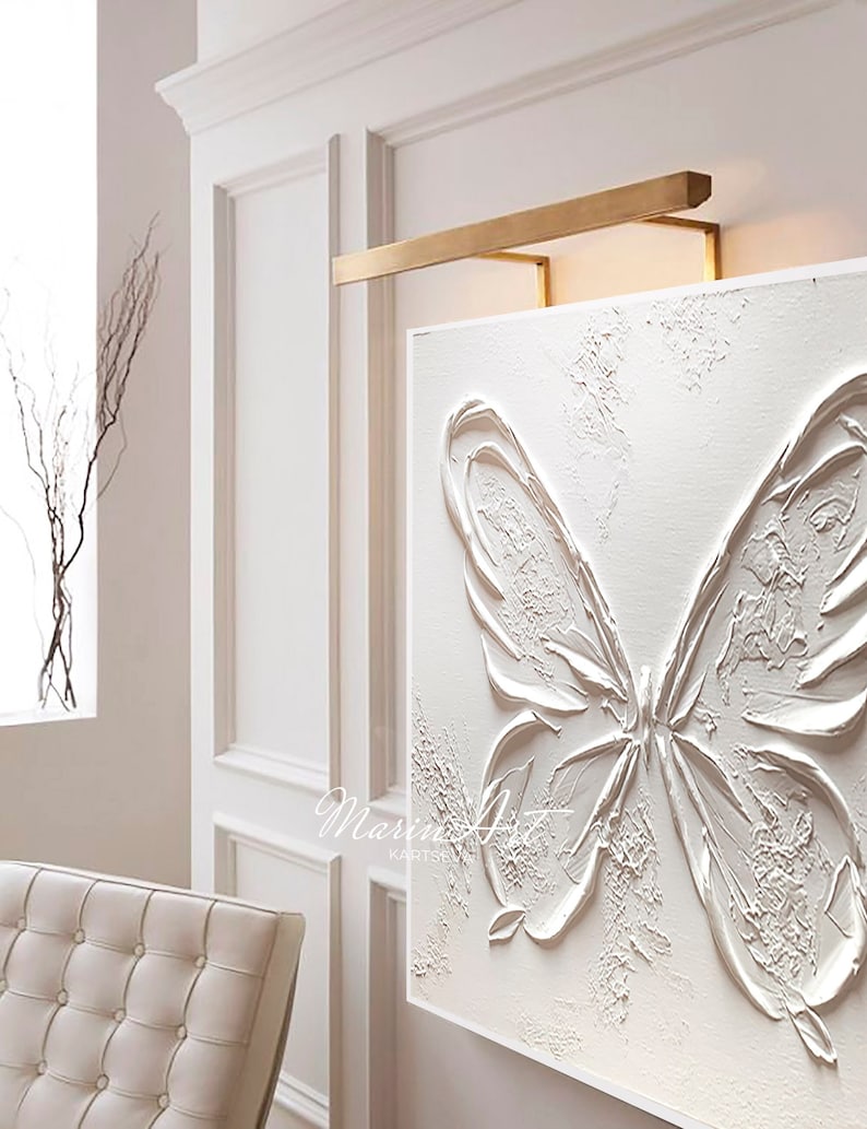 Op de afbeelding: Witte textuurverf op canvas van een vlinder met een gouden metalen frame en een lamp erboven. Het schilderij is getiteld "Marin Art".