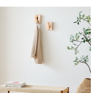 May include: A wooden wall mounted coat rack with two hooks. A beige sweater hangs on one of the hooks. A wooden bench with a woven seat sits in front of the rack. A potted plant with green leaves sits on the floor next to the bench.