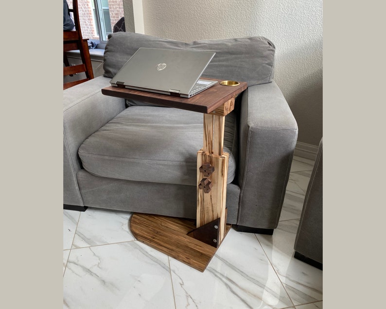 May include: A wooden laptop stand with a built-in cup holder. The stand is made of light and dark wood and has a metal accent. The stand is sitting on a gray couch.