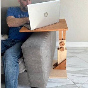 May include: A wooden laptop stand with adjustable height, featuring a natural wood finish and two wooden knobs. The stand is positioned on a gray couch, with a laptop resting on the top surface.