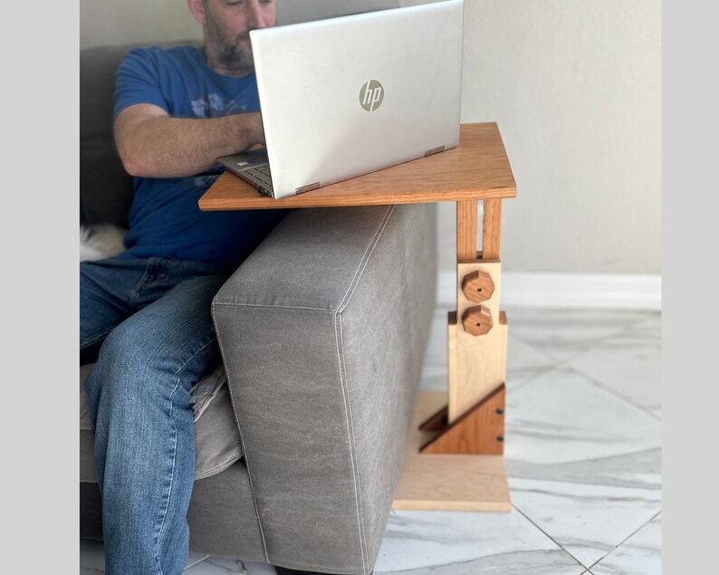 May include: A wooden laptop stand with adjustable height, featuring a natural wood finish and two wooden knobs. The stand is positioned on a gray couch, with a laptop resting on the top surface.