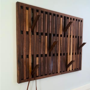 May include: A rectangular wall-mounted coat rack constructed from vertical wooden slats in varying shades of dark and light brown. The rack features several angled wooden hooks extending outward from the slatted frame for hanging items.