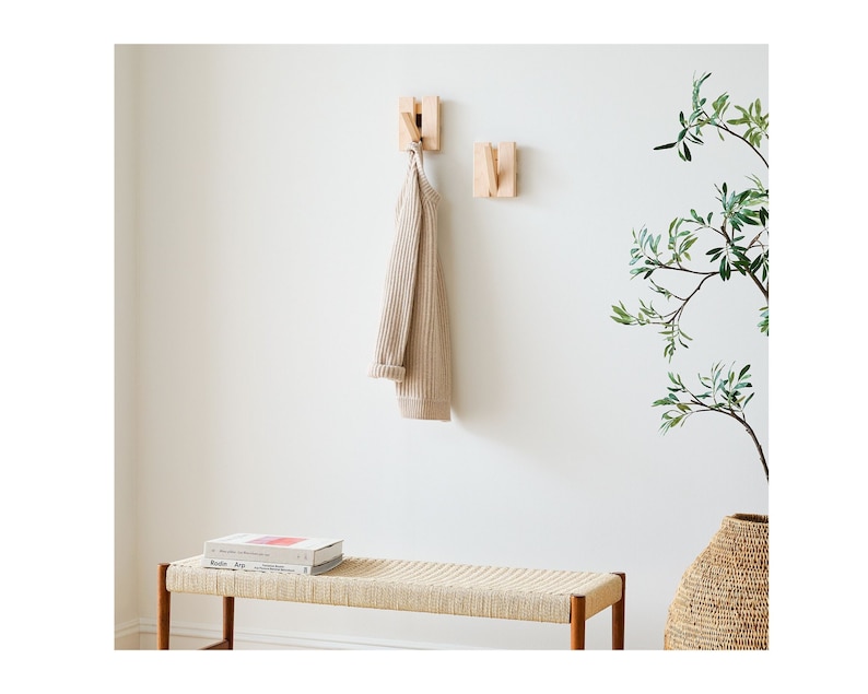May include: A wooden wall mounted coat rack with two hooks. A beige sweater hangs on one of the hooks. A wooden bench with a woven seat sits in front of the rack. A potted plant with green leaves sits on the floor next to the bench.