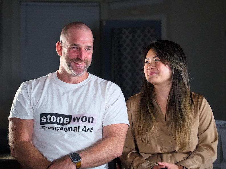 以下が含まれることがあります： 男性と女性が笑顔で向き合っています。男性は「stone won Functional Art」と書かれた白いTシャツを着ています。女性は薄い茶色のシャツを着ています。