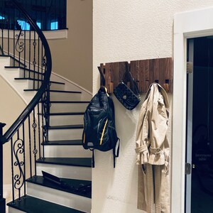 May include: A wooden wall-mounted coat rack with a dark brown finish. A black backpack, a brown leather purse, and a beige trench coat are hanging on the rack. The rack is mounted on a white wall.
