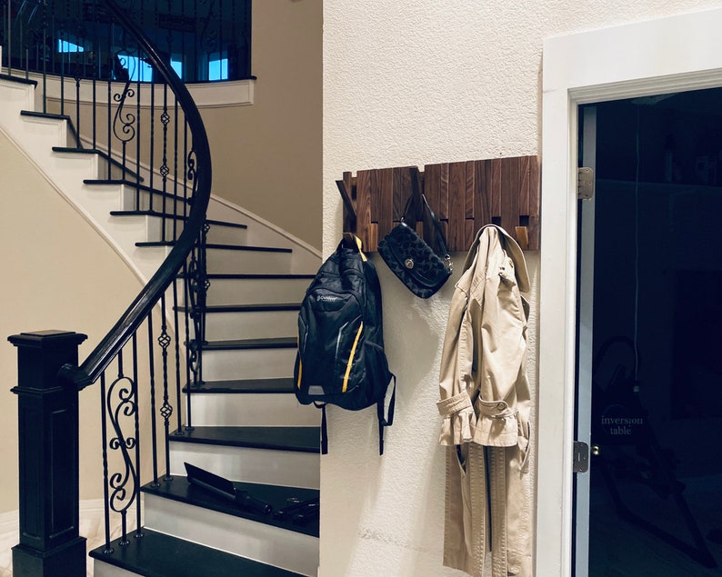May include: A wooden wall-mounted coat rack with a dark brown finish. A black backpack, a brown leather purse, and a beige trench coat are hanging on the rack. The rack is mounted on a white wall.