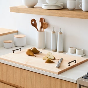 May include: A wooden cutting board with two handles, a knife, and sliced pears on a white countertop. The cutting board is made of light wood and has a natural finish. The knife is stainless steel and has a black handle. The pears are ripe and have a yellow skin. The countertop is white and has a smooth surface.