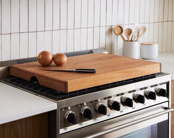 May include: A wooden cutting board sits on top of a stainless steel stovetop. The board has a lip around the edge to prevent items from sliding off. Two onions are on the board, along with a knife.