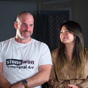 May include: A man and a woman are smiling at each other. The man is wearing a white t-shirt with the text "stone won Functional Art". The woman is wearing a light brown shirt.