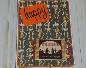 May include: A Halloween card with a gray background featuring a pattern of skeletons. The card has a bright orange border and a rectangular orange sticker with the word "happy" written in black. The card also features a square image of a moonlit Halloween scene with the text "Halloween" and "Trick or Treat".