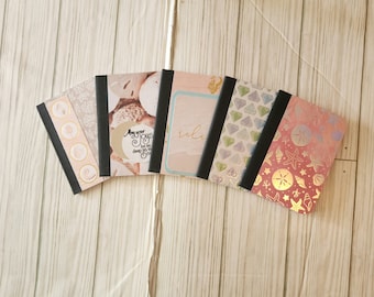 May include: Six small notebooks with different designs. The covers feature seashells, diamonds, and a watercolor-style background. The notebooks have black spines and gold lettering. The text on one notebook reads "May your joy be as deep as the sea".