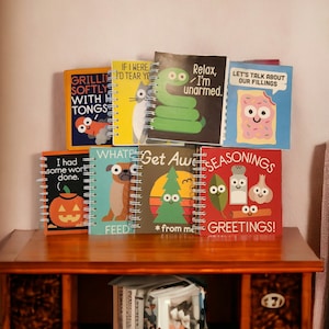 May include: A collection of eight colorful spiral bound notebooks with various illustrations and text. The notebooks feature a cat, a snake, a slice of toast, a pumpkin, a pug, a Christmas tree, a salt shaker, and a cinnamon stick. The text on the notebooks includes "If I were you I'd tear you", "Relax, I'm unarmed", "Let's talk about our fillings", "I had some work done", "What's for dinner", "Get Awesome", "Seasonings", "Feed me", "From me", and "Greetings!"
