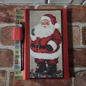May include: Red notebook with a Santa Claus illustration on the front cover. The notebook has a pen holder on the side and a red pen is inserted in the holder. The notebook has a patterned design on the back cover.