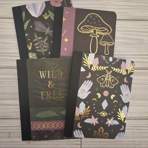 May include: Four black and gold notebooks with different designs. The first notebook has a crystal, leaves, and a butterfly design. The second notebook has a mushroom design. The third notebook has a "Wild & Free" text design. The fourth notebook has a hand, eye, crystal, and moth design.