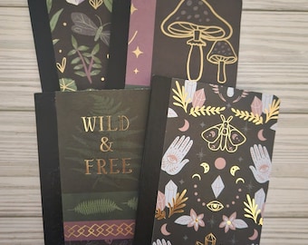 May include: Four black and gold notebooks with different designs. The first notebook has a crystal, leaves, and a butterfly design. The second notebook has a mushroom design. The third notebook has a "Wild & Free" text design. The fourth notebook has a hand, eye, crystal, and moth design.