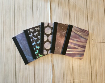 May include: Five small notebooks with different designs. The notebooks have black covers and are stacked on a wooden surface. The designs include a moon and stars, a galaxy, a unicorn, and a marble pattern.