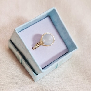 May include: A white gemstone ring with a gold wire wrap setting, displayed in a light blue ring box.
