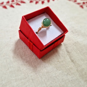 May include: A gold ring with a green gemstone set in a red ring box.