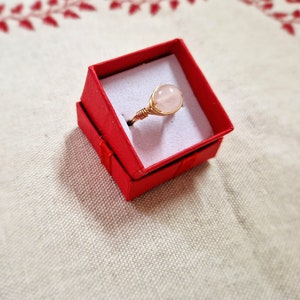 May include: A delicate ring with a light pink gemstone set in a gold wire wrapped band. The ring is displayed in a red velvet ring box.