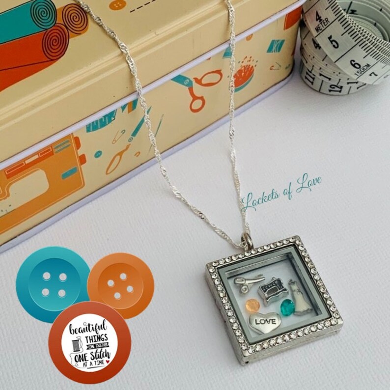 May include: A silver locket necklace with a square frame and clear glass insert. The locket is filled with small charms, including a sewing machine, scissors, a button, a heart with the word "LOVE" on it, and a turquoise gem. The necklace is on a white background with a sewing-themed box and a measuring tape.