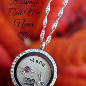 May include: A silver locket necklace with a clear crystal encrusted bezel. The locket has a silver background with the word "Nana" engraved on it. Inside the locket are three charms: a sewing machine, a pair of scissors, and a ball of yarn with needles. The necklace is on a red and orange floral background. The text "My Greatest Blessings Call Me Nana" is written above the locket. The text "Lockets of Love" is written below the locket.
