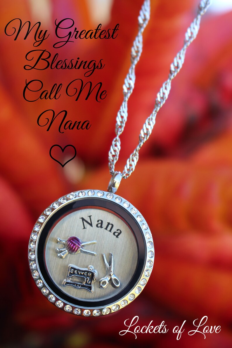 May include: A silver locket necklace with a clear crystal encrusted bezel. The locket has a silver background with the word "Nana" engraved on it. Inside the locket are three charms: a sewing machine, a pair of scissors, and a ball of yarn with needles. The necklace is on a red and orange floral background. The text "My Greatest Blessings Call Me Nana" is written above the locket. The text "Lockets of Love" is written below the locket.