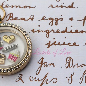May include: Gold locket necklace with a clear glass insert and a crystal-encrusted border. Inside the locket are charms including a pink mixer, a rolling pin, a cupcake, and a heart charm with the word "NANA" on it.