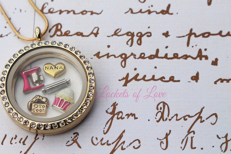 May include: Gold locket necklace with a clear glass insert and a crystal-encrusted border. Inside the locket are charms including a pink mixer, a rolling pin, a cupcake, and a heart charm with the word "NANA" on it.