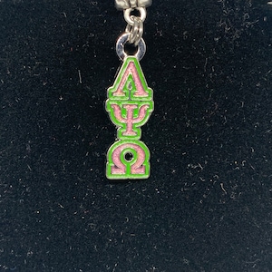May include: A green and pink charm with Greek letters hangs from a silver pin. The charm is shaped like a stylized column and is displayed on a black velvet bust.