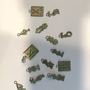 May include: A selection of silver charms with diverse designs, including letters, symbols, and geometric shapes. Some charms have pink and green details. The charms are arranged on a white surface.
