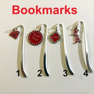 May include: Four silver bookmark holders with different charms. The charms are a Superman logo, a red circle with the words "Delta Dear" in white, a clover with the letters "ΔΣΘ" in white, and a red figure of a woman with the letters "ΔΣΘ" in white.