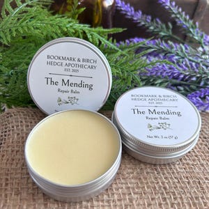 May include: Two silver tins of "The Mending Repair Balm" by Bookmark & Birch Hedge Apothecary, one open, revealing a light yellow balm. The tins are on a burlap surface, with green and purple foliage in the background.