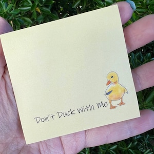 Könnte beinhalten: Ein hellgelber Haftzettel mit dem Schriftzug "Don't Duck With Me" in schwarzer Schrift. Eine Cartoon-Entenküken-Illustration befindet sich in der Ecke. Der Zettel wird in einer Hand vor einem Hintergrund aus grünem Laub gehalten.