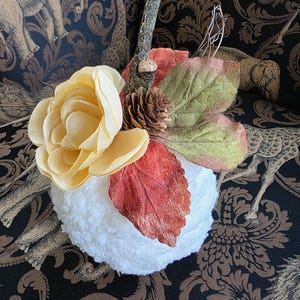 May include: A white textured fabric pumpkin decoration topped with a large pale yellow fabric flower, a small brown pinecone, a realistic twig stem, and several artificial autumn-colored leaves in shades of green, orange, and deep red.