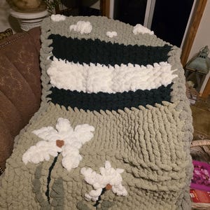 May include: A chunky knit blanket featuring a sage green base with horizontal stripes in dark green and white. The design includes white floral motifs with brown centers and small white cloud-like shapes at the top, all crafted from thick yarn.