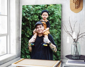 May include: A father and son are standing in a forest. The father is wearing a black shirt with a tattoo on his arm. The son is wearing a brown shirt and a black hat. They are both smiling.